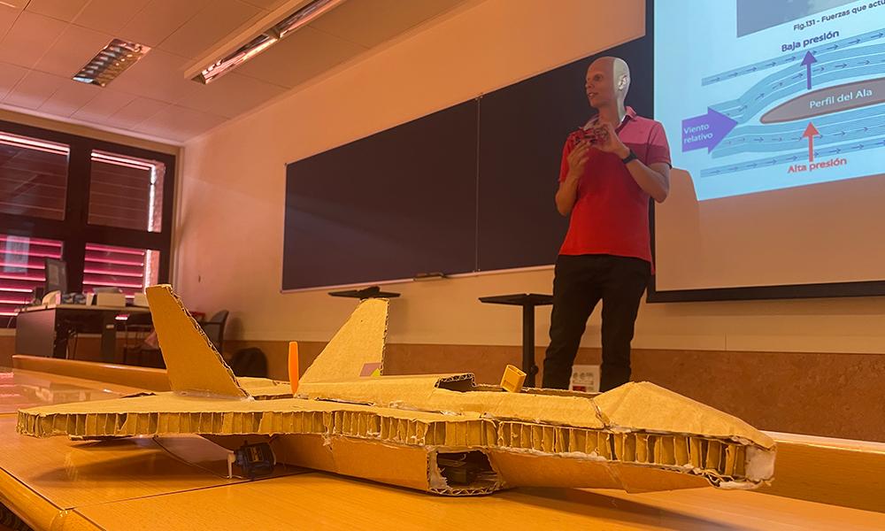 El profesor al fondo en la pizarra y delante una maqueta de avion