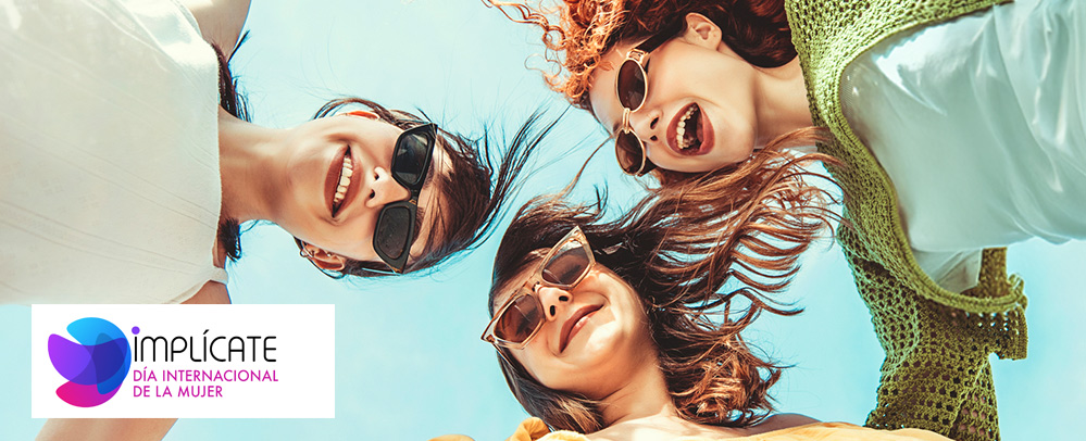 Tres mujeres jóvenes sonriendo juntas bajo un cielo despejado, vistas desde un ángulo contrapicado.